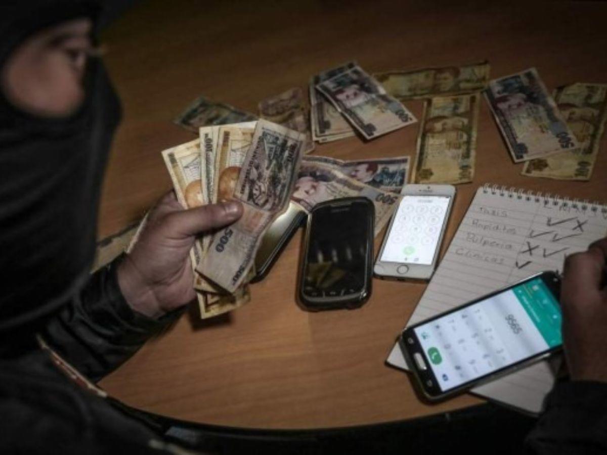 hombre con capucha contando dinero y con celulares sobre una mesa