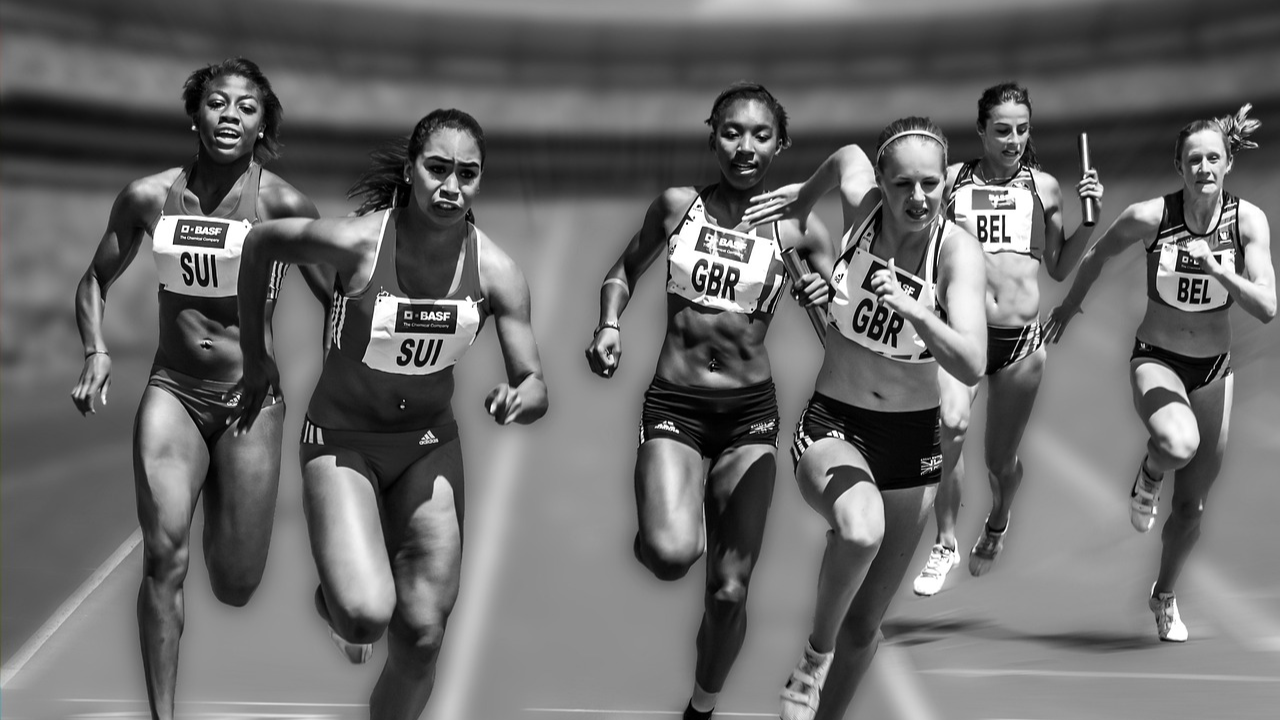 Imagen es blanco y negro de 6 mujeres jóvenes corriendo por una pista de atletismo. 