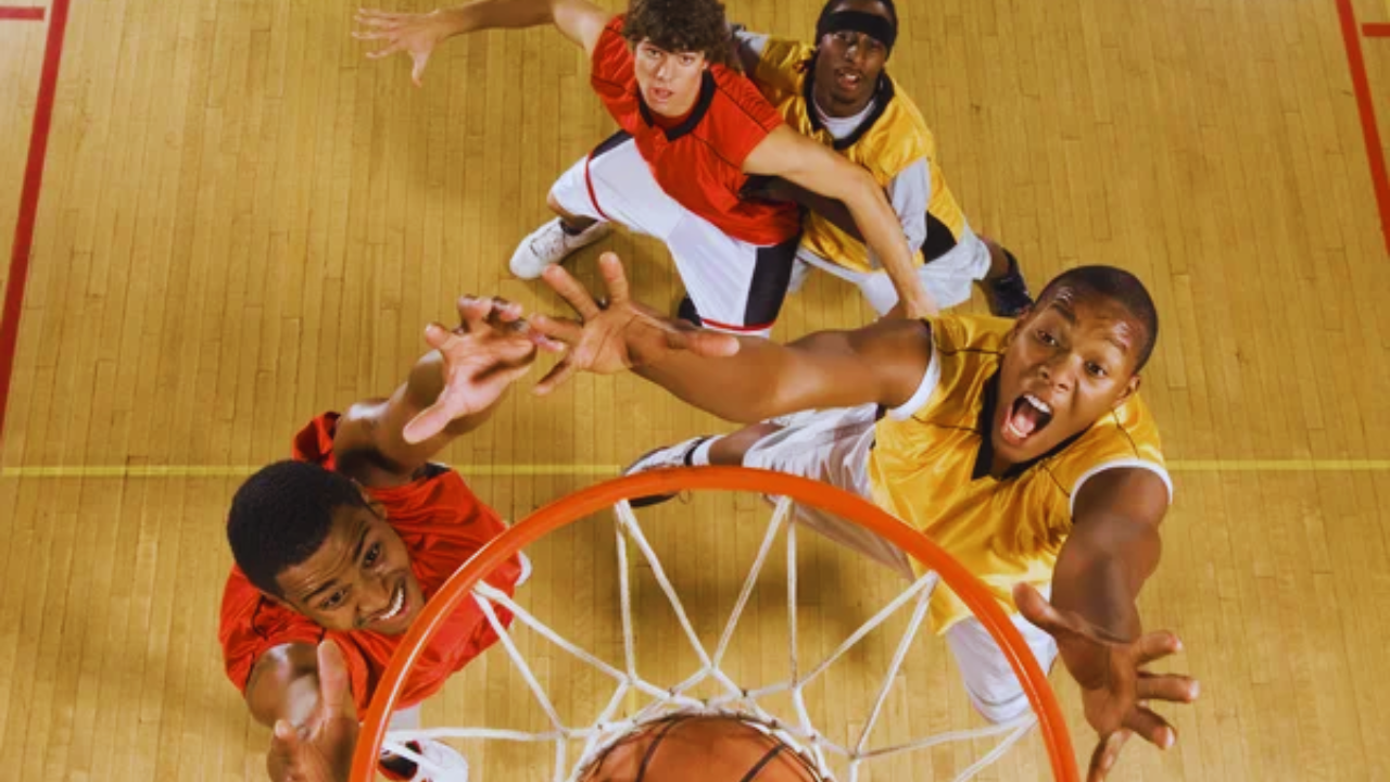 Una cancha de baloncesto color café claro y 4 hombres jugando baloncesto con un balón anaranjado con rayas negras. 