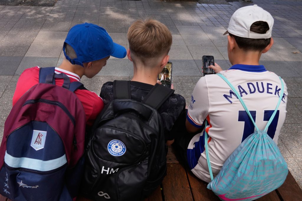 Hugo Winwood-Smith, a la derecha, Hardy Macpherson y Edan Abou, a la izquierda, todo de 11 años, utilizan sus celulares mientras se sientan ante una escuela en Sydney, el lunes 8 de diciembre de 2025. (AP Foto/Rick Rycroft)