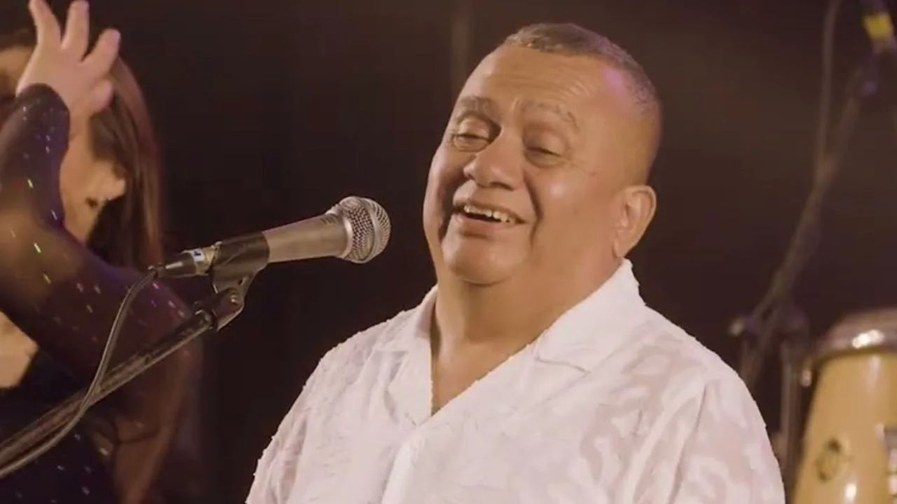 Tony Rosado con una camisa blanca cantando frente a un micrófono.