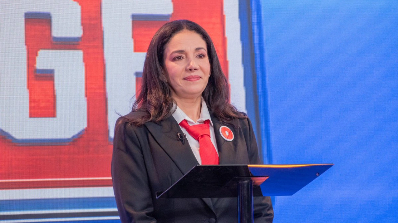 Sondra Macollins frente a un atril vestida de saco negro, corbata roja y con camisa blanca.