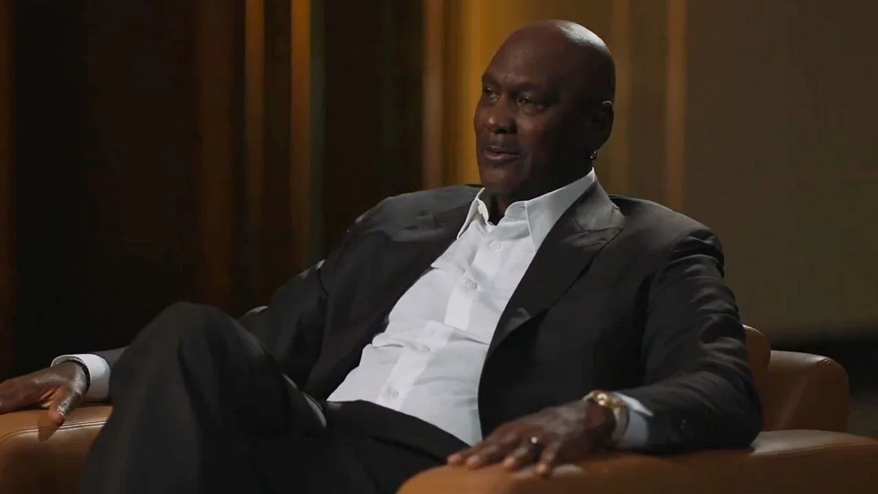 Michael Jordan sentado y vestido con un traje negro y camisa blanca de botones.